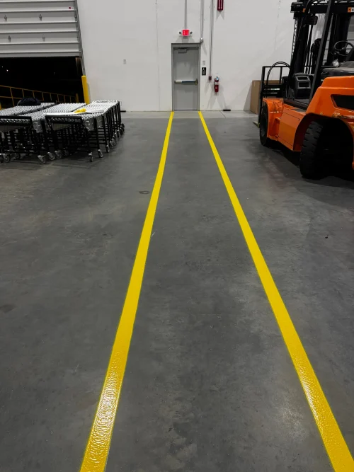 A warehouse with a bright orange forklift on the right, several rows of carts on the left, and a gray door with an exit sign in the back. Two parallel yellow lines run down the concrete floor in the middle.